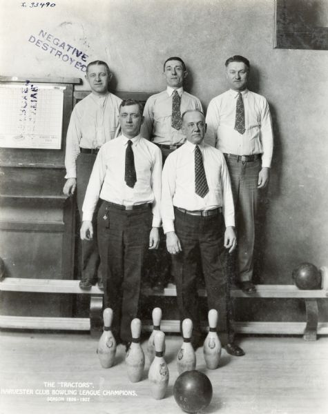 Five men standing behind a bowling ball and pin display for a gropu portrait. The men are members of the "Tractors" bowling team, Harvester Club Bowling League champions for the 1926-1927 season.