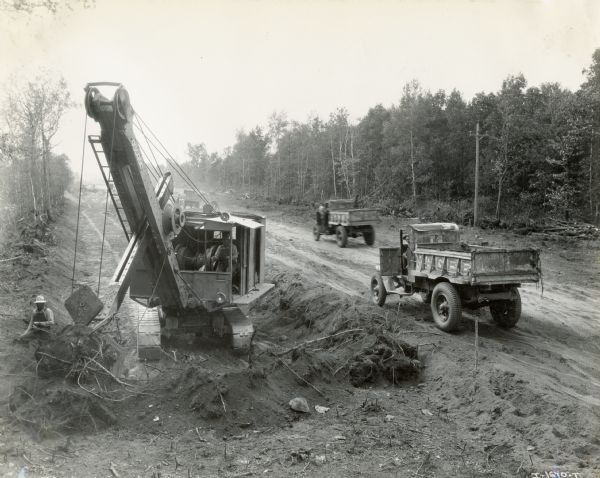 Road construction crews use an excavator to dig while two dump trucks drive by in the background. The trucks are likely Internationals.