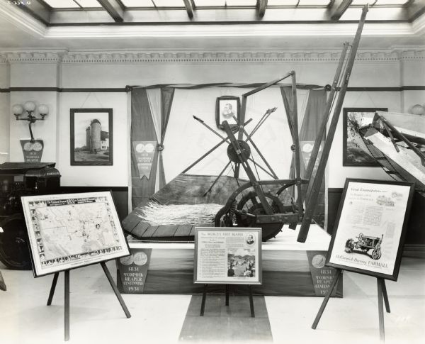 A replica of Cyrus McCormick's first reaper on display with signs and posters. The display was likely part of the "reaper centennial," a promotional event for the International Harvester Company.