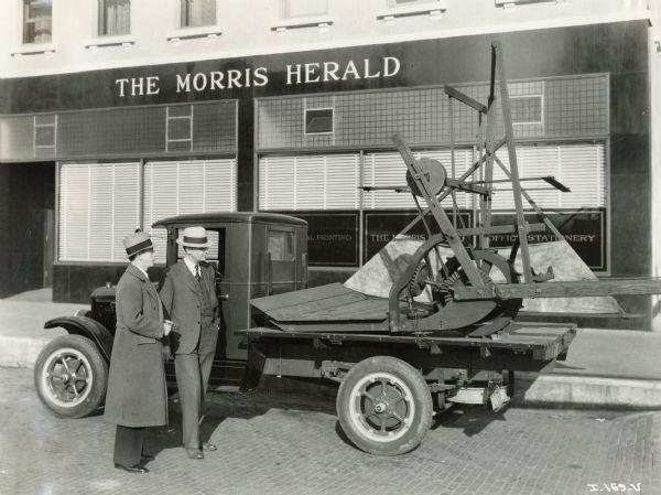Two men examine a replica of McCormick's original reaper on the bed of an International Six-Speed Special truck. The replicas were produced for the "reaper centennial celebration," a promotional event for International Harvester. The truck is parked in front of the "Morris Herald" building.