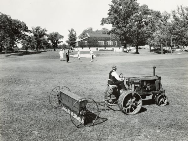 A groundskeeper uses a McCormick-Deering Fairway tractor to care for the golf course at Indian Head Golf Club. A group of people are playing golf in the background.