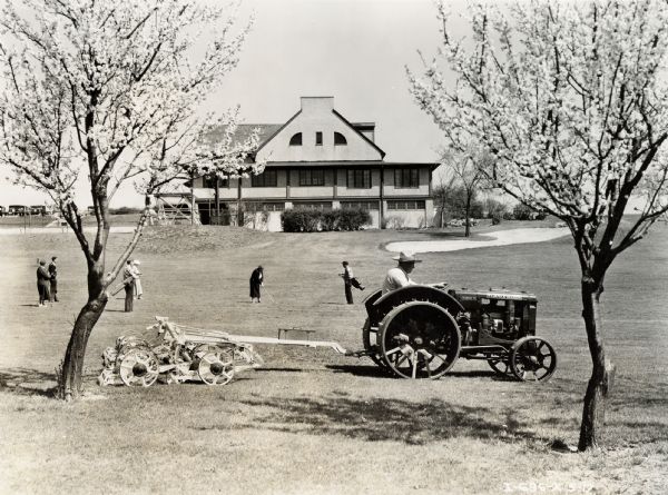 A man uses a McCormick-Deering Fairway 12 tractor and a mowing attachment to maintain a golf course. A group of people are playing golf in the background.