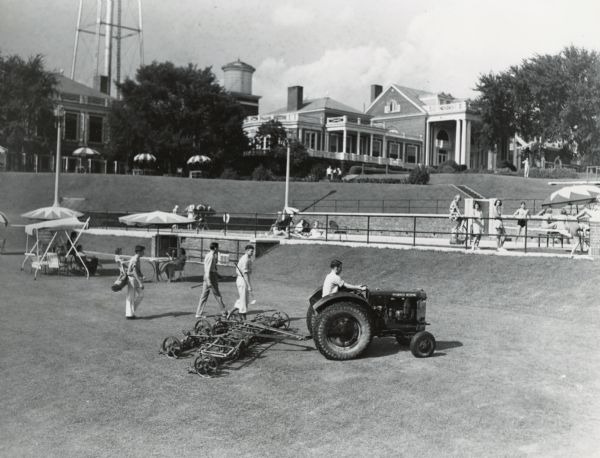 Man uses a McCormick-Deering Fairway 12 tractor and mowing attachment to care for lawns (and golf course?) at a country club. Sun bathers are in the background.