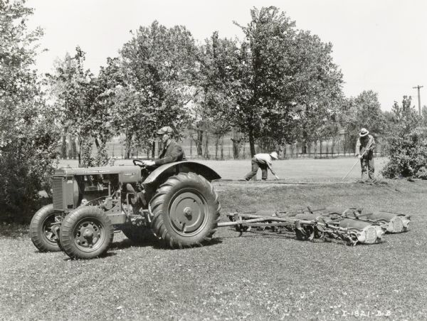 A man uses a rubber-tired Fairway 12 tractor with a Roseman mower attachment to care for golf course terrain. Two men are working in the background.