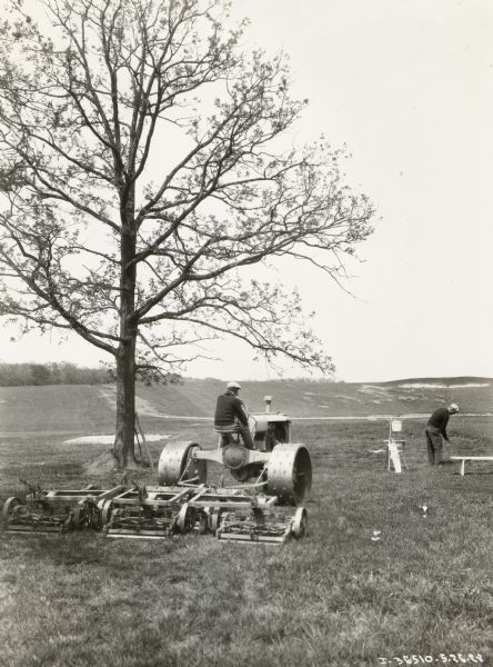 A man on a Fairway tractor with a mower waits while a golfer tees off at a golf course.