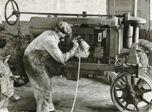 A man wearing coveralls and a mask spraying paint(?) on a Farmall  tractor. The man is probably a service technician or mechanic at an Interational Harvester dealership.