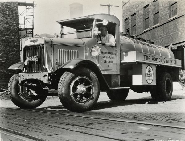 Driver sitting in an International truck operated by the Mobiloil Company. Text on the side of the truck reads: "The World's Quality Oil."