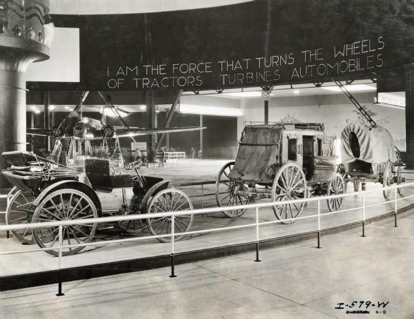 A covered wagon, horse-drawn carriage (coach), and an automobile on display at the "A Century of Progress" world's fair in Chicago. Sign above the display reads: "I am the force that turns the wheels of tractors, turbines, automobiles."