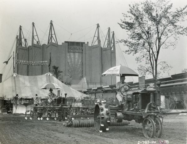 Two men are operating Farmall tractors and implements at the "A Century of Progress" world's fair. One tractor is pulling a disk harrow, and the other a cultivator. An International Harvester Company tent and the Travel and Transport Building are in the background.
