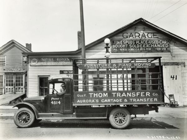 The driver of an International Model S truck sits in the cab outside of a building marked "Garage" with a sign underneath reading: "Jim and Spuds Place - Used Cars Bought, Sold and Exchanged; Small Payment Down and $5 a Week." Two other signs on the right part of the building over a small window read: "Spire Bros. Used Cars Bought, Sold and Exchanged" and "Gus F. Thom Transfer Co." The truck is labeled with the text "Piano Moving; Finest Equipment Expert Moving; Gus F. Thom Transfer Co.; Aurora's Cartage and Transfer."