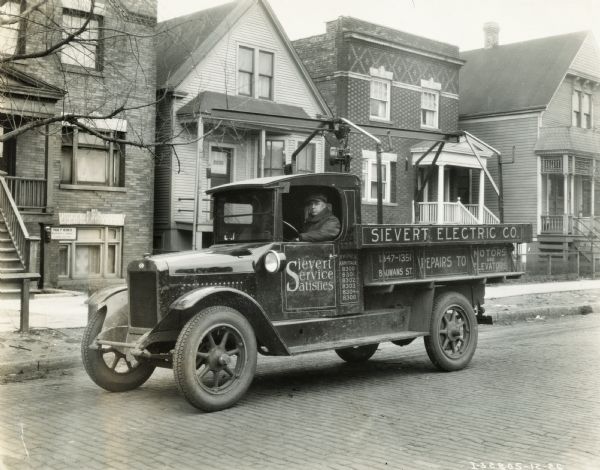 International Model S truck parked on a residential street. The truck was operated by the Sievert Electric Company. Text on the side of the truck reads: "Sievert Service Satisfies; Sievert Electric Co.; Repairs to Motors and Elevators."