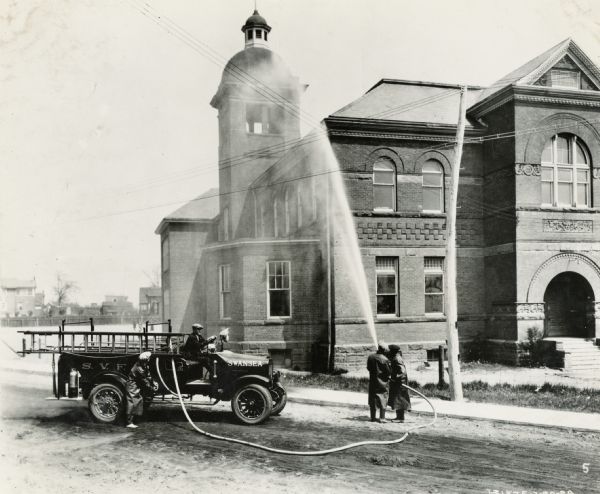 Two men are using a fire hose to spray the corner of a brick building with a bell tower. Another man is sitting in an International Model S truck operated by the Swansea Fire Department.