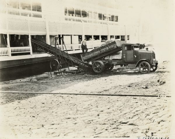 A man shovels coal from the bed of an International Model 63 truck onto a conveyor attached to a river boat or steam boat. The truck was used by the "Thos. J. Mulgrew Company" to haul "Coal Coke [and] Ice". Cooks, a kitchen, and a rocking chair are on the boat.