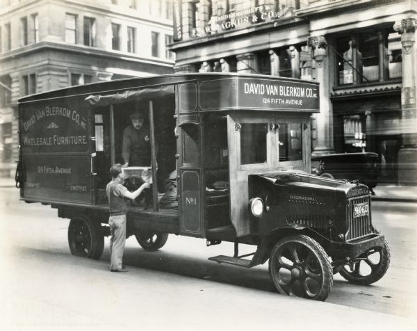 Two men unloading from the back of an International Model 63 truck on a city street. The truck was owned and operated by the David Van Blerkom Company, dealers in "wholesale furniture."
