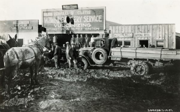 International Model 63 truck loaded with lumber, and parked in a muddy street outside the "Ishmael and Curtis Willard Battery Service" building. A sign on the building includes the text "Vulcanizing Automotive and Oil Field Ignition." Horses and other people are standing on the street and on a boardwalk. Other buildings on the street include Kernes Cafe "Meals Family Style 45 cents," Model Studio "Kodak Finishing Specialty" and the Ramona Hotel. A small sign above the Willard store sign reads "Miller Tires".