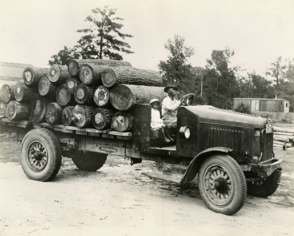 A man and a young girl (daughter?) sitting in the open cab of an International Model 63 truck loaded with logs.