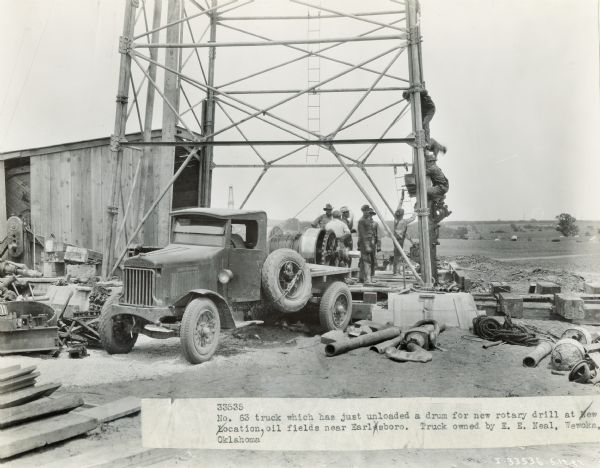Men set up a drum for a new rotary drill that has just been unloaded from an International Model 63 truck belonging to E.E. Neal of Wewoka, Oklahoma. The oil field was near Earlsboro. Oil drilling machinery and tools are on the ground.