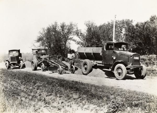 A man stands on a road surrounded by two International Model 63 trucks and a Gilbert road grader machine operated by the M.O. Weaver Construction Co.