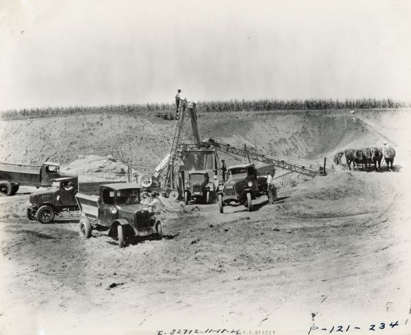 Elevated view of men and a team of four horses working among dump trucks, including an International Model 63, at a Moore and Eldredge construction site.