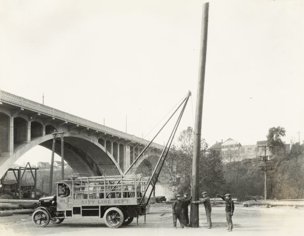 Workmen with the City Line Department use an International Model 63 truck to erect a power line or telephone pole near a bridge.