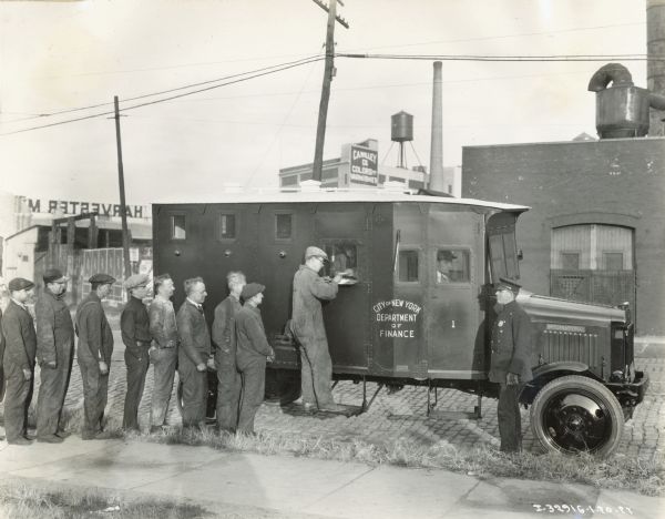 A group of men line up for their pay at an armored International Model 63 truck used by the City of New York Department of Finance. The truck is guarded by a security guard or police officer.