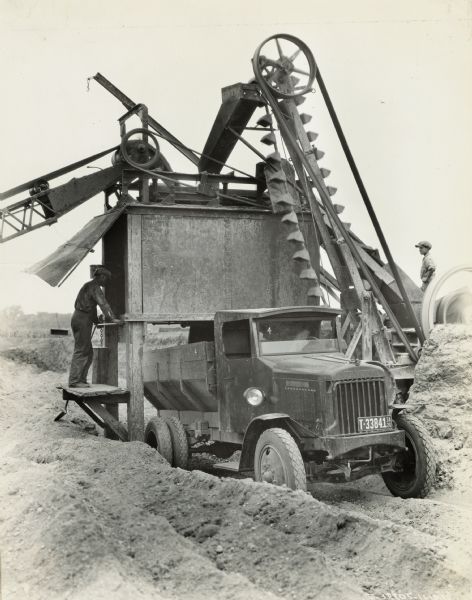 A man and a young boy stand on either side of an International Model 63 dump truck as it is loaded. The truck was operated by the M.O. Weaver Construction Company.