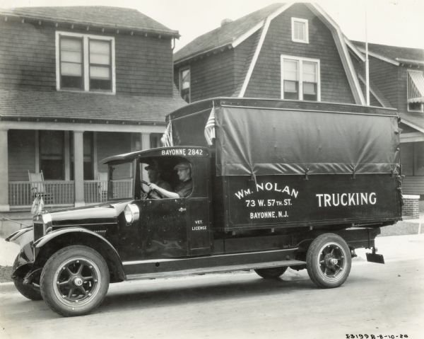 Two men are sitting in the cab of an International Model S truck used by William Nolan Trucking. There are two small American flags behind the roof of the cab, and more flags are mounted on the front of the hood. The truck is on a residential street, with houses in the background. The signs painted on the side of the truck read: "Bayonne 2842," "Vet. License," and "Wm. Nolan, Trucking."