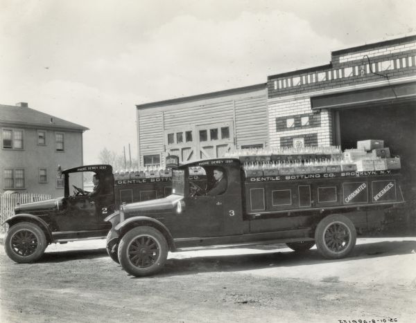 Two men are posing sitting in the driver's seats of two International Model S trucks loaded with "carbonated beverages" from the Gentile Bottling Company of Brooklyn.