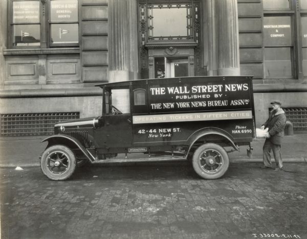 A man loads newspapers onto an International Model S truck used by "The Wall Street News". Text on the truck reads: "The Wall Street News; Published by the New York News Bureau Assn. Inc. Operating tickers in fifteen cities. 42-44 New St. New York. Phone Han.6990." The truck is parked on a city street in front of a building that includes Wightman, Breining & Company.