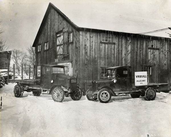 Two International Model S trucks with snow chains on their tires parked outside a barn. A sign on one of the trucks reads: "WM N. Hall, Allegany, NY."