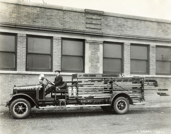 A man is sitting behind the wheel of an International Model SF 1926 fire truck used operated by the Center Moriches Fire Department. The text on the truck reads: "Center Moriches, F.D., H&L No.1."
