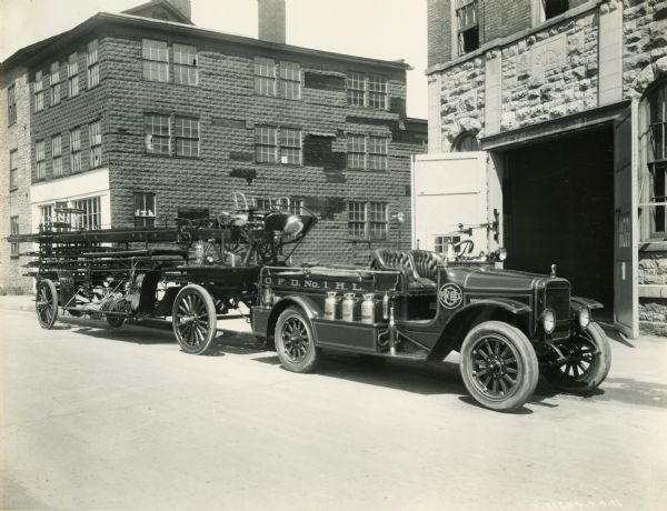 An International Model S truck used as a fire truck and attached to a trailer holding a long, folded ladder sits parked outside Engine House No. 2. The sign on the fire engine reads: "O.F.D. No.1 H.L."