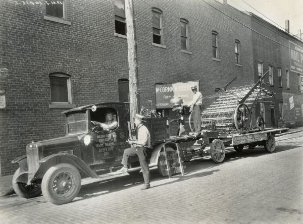 International Model S truck operated by the Bluffton Implement Company, an International Harvester dealer. A man is standing with his foot on the running board while another man is sitting at the wheel. A trailer with a hay loader and cream separator is attached to the truck. The sign on the building behind the truck reads: "McCormick-Deering Farm Machines; Bluffton Implements".  The owner of the dealership was Ware Baker.