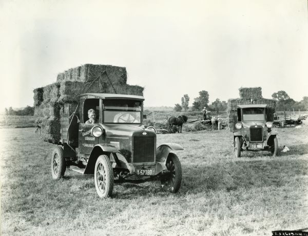 A man and a young boy are sitting in the cab of an International Model S truck loaded with bales of hay as men with horses are working in the background. The text on the door of the truck reads, in part: "McCormick-Deering; Gard... Graham; Carlisle."