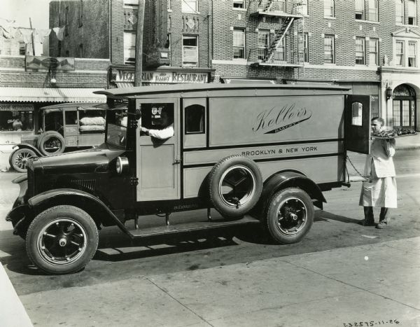 A driver sits behind the wheel of an International Model S truck used by Keller's Bakery of Brooklyn as a man loads a tray of goods into the back. The storefront signs in the background read: "Boot Black Parlor and Hat Cleaning" and "Vegetarian Dairy Restaurant".