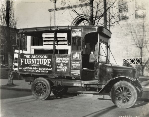 An International Model S truck used by the Jackson Furniture House  parked alongside a curb with its rear compartment full of wooden pallets. The text on the truck reads: "The Jackson Furniture House, 237 Jackson Ave. near Bayview Ave. Jersey City, New Jersey. Dining Room, Bed Room, & Parlor Suites; Complete Line of House Furniture; Sheinkin Brothers, Props."