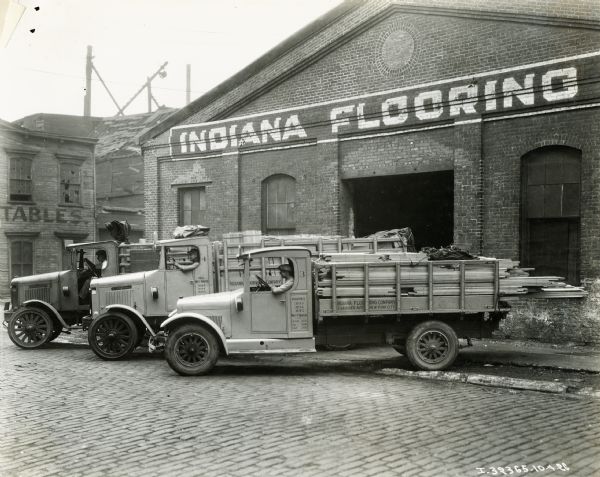 Three men sitting in the cabs of International trucks loaded with lumber. They are sitting in a row outside of what appears to be a warehouse of the Indiana Flooring Company. At least one of the trucks is an International Model S. The text on one of the trucks reads: "Indiana Flooring Company, 234 Rider Ave., New York City."