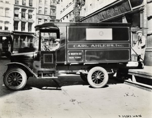 A man loads a box onto the back of an International Harvester Model S truck used by Carl Ahlers, Incorporated. Another man sits in the cab of the truck.