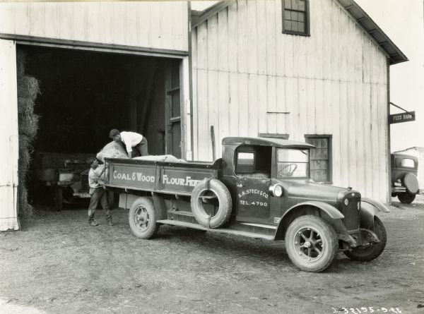 Two men unload (or load) bags from the back of an International Model S truck used by S.A. Steck & Co. A sign on side of building reads: "Feed Barn". The text on the truck reads: "Coal & Wood, Flour, Feed...(?)"