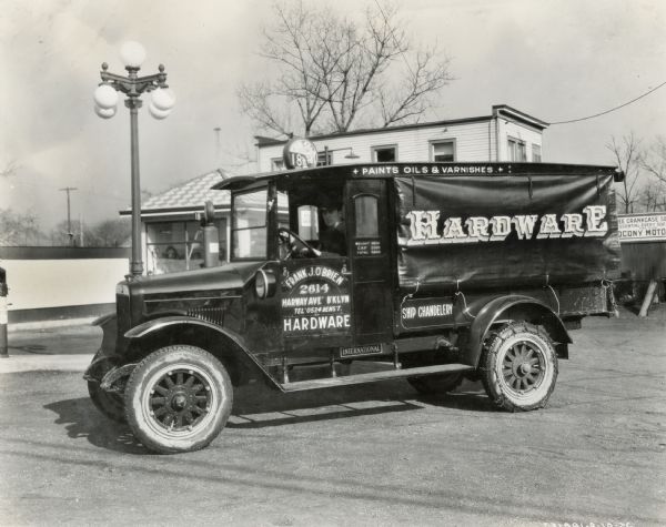 A driver sits behind the wheel of an International Model S truck used by the Frank J. O'Brien Hardware Company. A service station is in the background. The text on the truck reads: "Frank J. O'Brien, 2614 Harway Ave. B'kyln; Hardware; Paints, Oils, & Varnishes; Ship Chandelery."