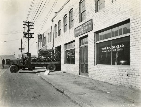 An International truck carrying a Farmall Regular tractor backs into a garage at the Evans Implement Company, an International Harvester dealership. The text in the windows of the store reads: "McCormick-Deering Tractors & Equipment" and "Meadows, Mills, Wehr Graders".