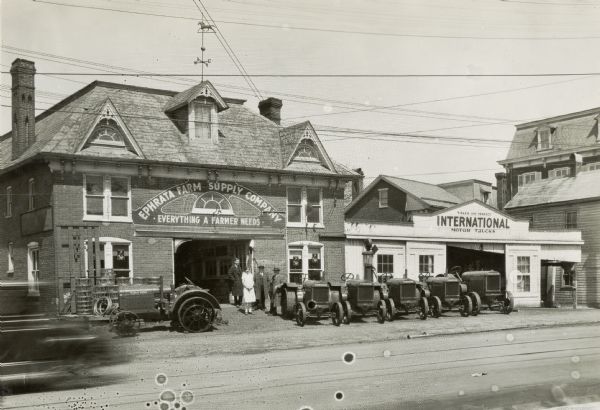 Several people are standing near the doors outside the Ephrata Farm Supply Company, an International Harvester dealership. Tractors are lined up outside the building. Signs on the dealership building read: "Everything a Farmer Needs" and "Sales and Service; International Motor Trucks."