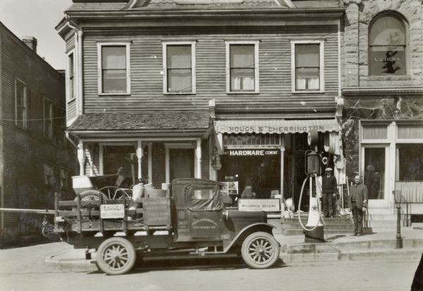 Two men stand on the steps leading into the Houck & Cherrington hardware store while an International sales and service truck is parked alongside the curb in front of them. The store may have been an International Harvester dealer.