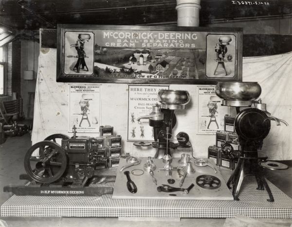 Ball-bearing cream separators, parts, and a one and half horse power stationary engine on display against a backdrop of advertising posters at the Milwaukee Works factory.