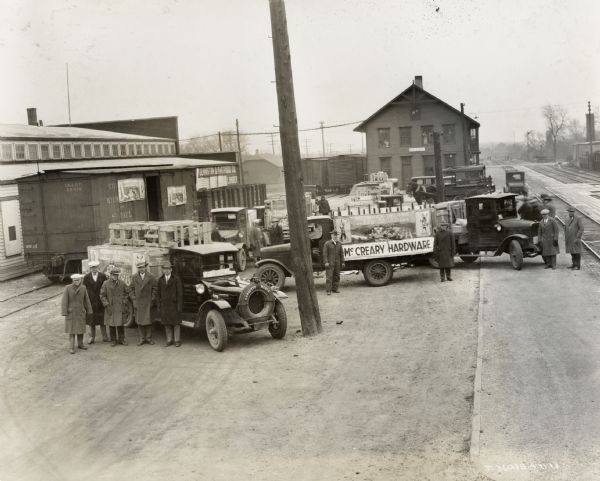 Men, most likely International Harvester dealers, and trucks gathered around a railroad boxcar with opened doors. The boxcar and a truck are adorned with posters advertising McCormick-Deering cream separators. The trucks are loaded with crates - presumably recently arrived cream separators. One of the trucks bears a sign reading: "McCreary's Hardware."