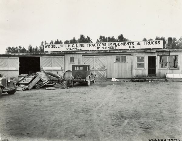 A man kneels to work on an automobile in front of the Chappell Implement Company, an International Harvester dealership. The sign above the building reads: "We Sell the I.H.C. Line - Tractors, Implements, & Trucks."
