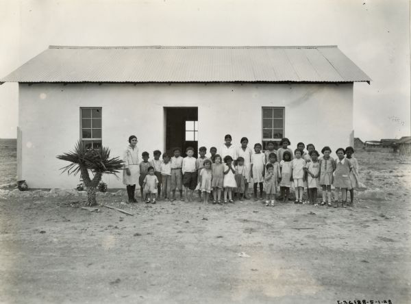 A group of Mexican school children pose with their teacher near a palm tree in front of a schoolhouse.