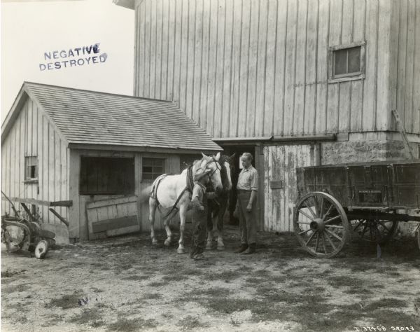 Two men stand beside a pair of horses outside a barn. A McCormick-Deering Weber wagon is on the right.