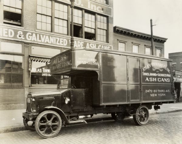 International Model 63 truck used by the Atlas Tinware Company parked in front of the company building. A blacksmith shop is in the background. Text on the side of the truck reads: "Atlas Tinware Co., Inc., Manufacturers of Tinned, Japanned, and Galvinized Ware Ash Cans."