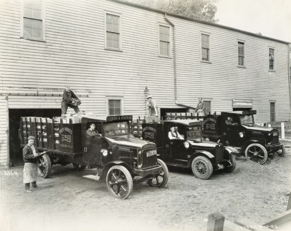 Elevated view of men posing with delivery trucks in front of the open garage door of a large, two-story wood building. Signs on the trucks read: "Quality Products F&H Brand, Newark, N.J.," and "Fountain & Heinzman." Three men are sitting in the passenger seats of the three trucks, one man is standing on the left and wearing an apron and holding a board, and three more men are each standing on top of boxes of bottles stacked in the truck beds. At least one of the trucks is an International Model 63.